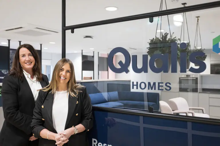 Two women smiling in an office next to Qualis Homes logo