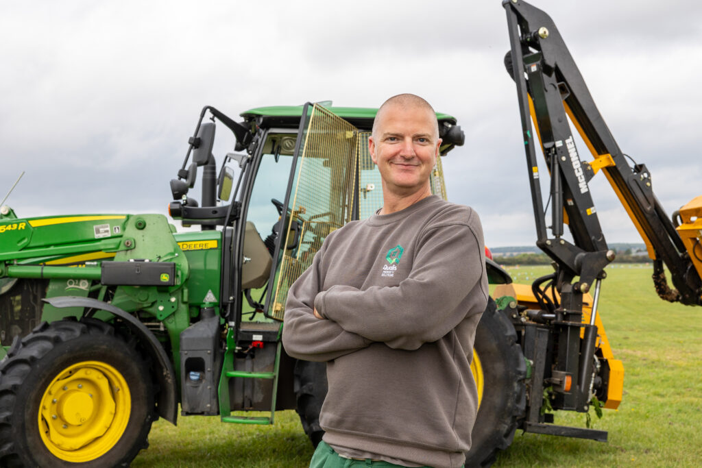 Man wearing Qualis branded sweatshirt stands in front of a tractor