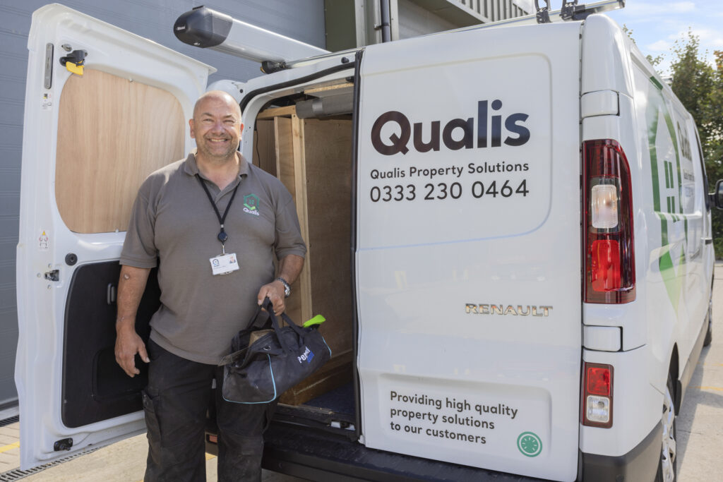 Man stands holding a tool bag at the door of a Qualis Property Solutions branded van.