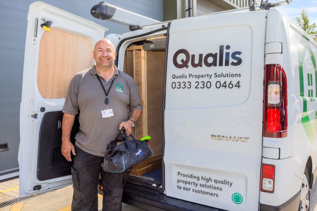 Handyman wearing Qualis branded polo shirt stands holding toolbox at the back of an open van.