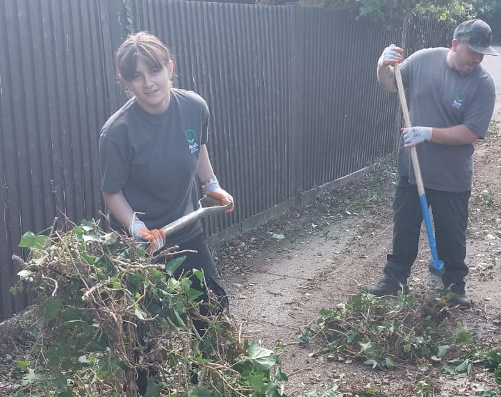 Young female stands with a shovel collecting garden waste. A second person is part visible in the background overseeing her work.