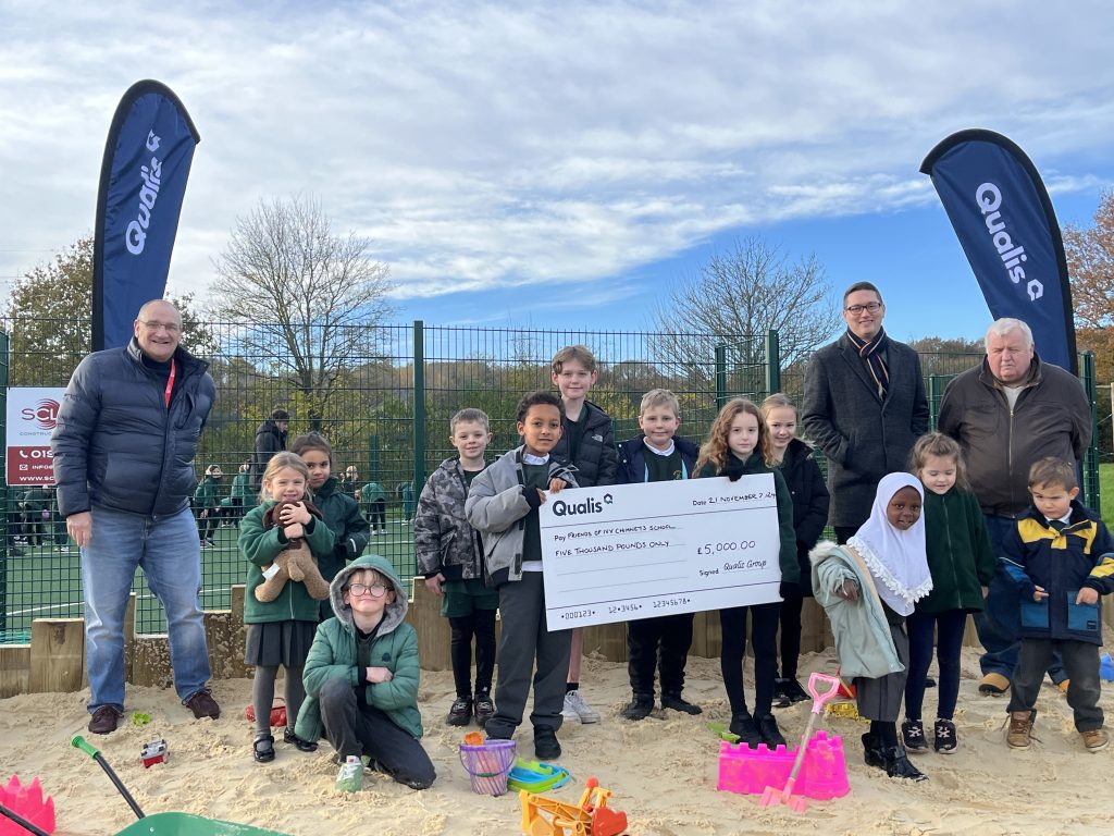 Group of children and adults stand in a sand pit holding a giant cheque
