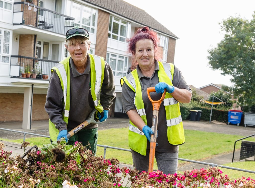 Two women stand in high vis jackets behind a flower bed. Both women carry gardening shovels.