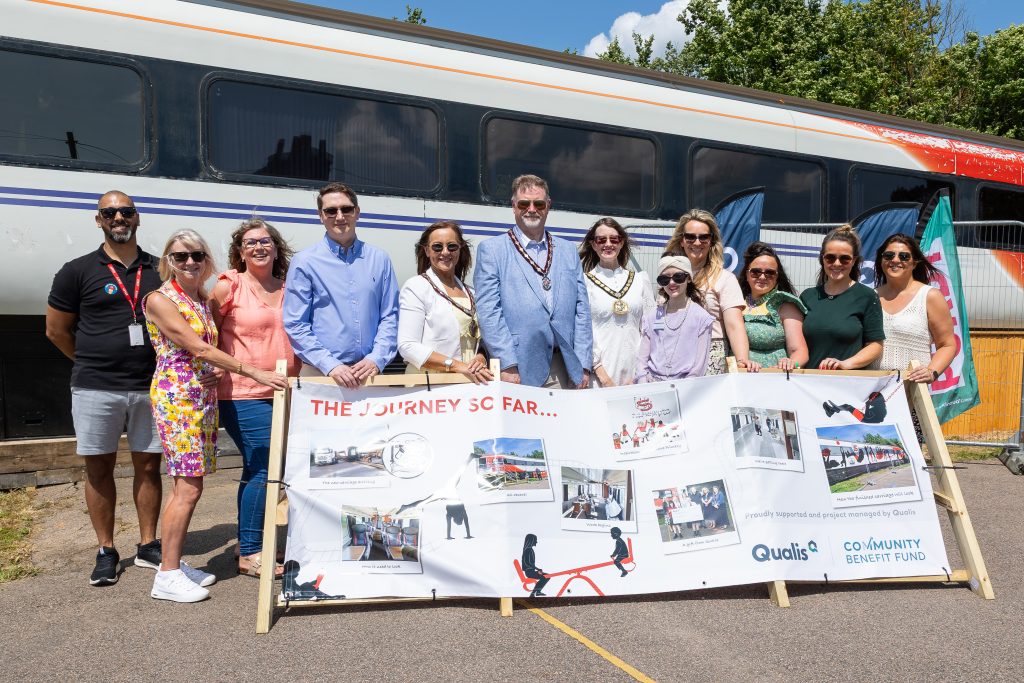 Group of people stand behind a banner and in front of a train carriage smiling.