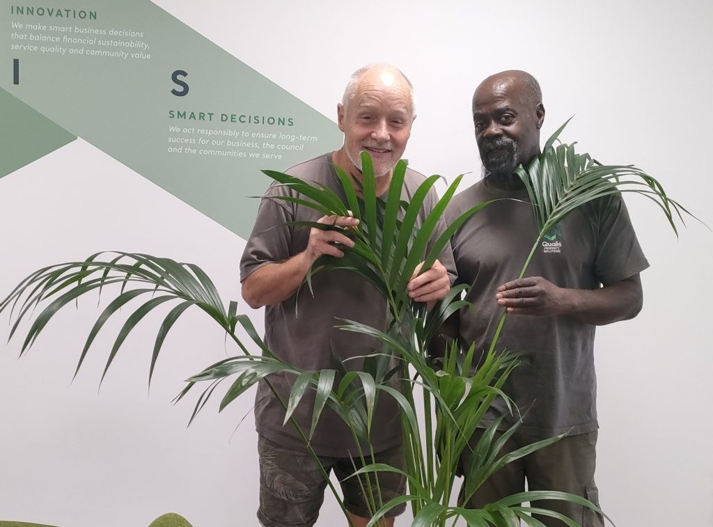Two men wearing Qualis uniform stand behind a pot plant